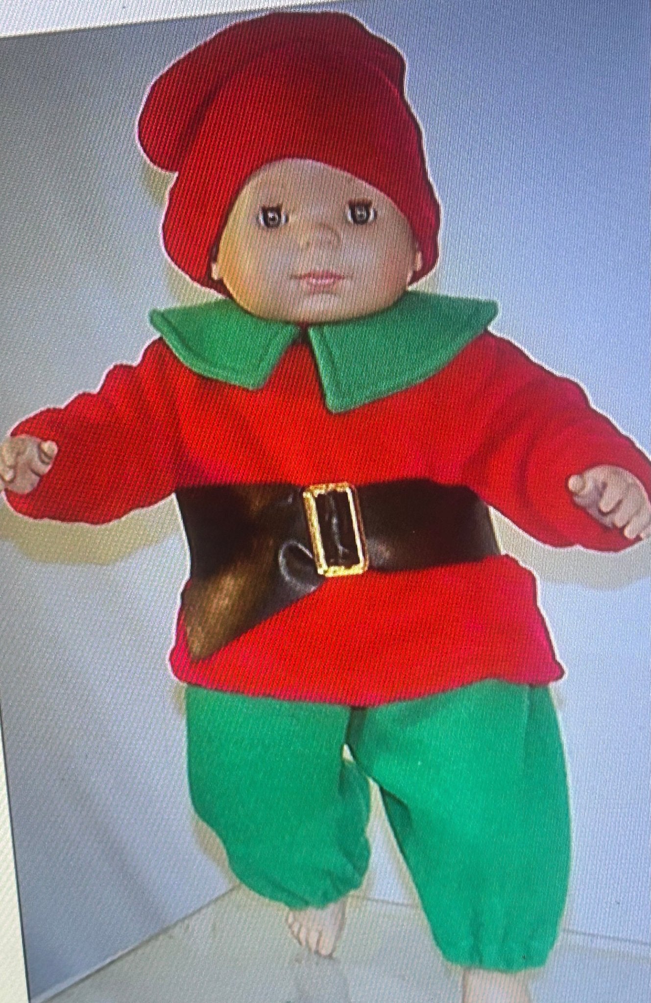Muñeco de bebé con traje rojo, capucha y pantalón verde, cinturón y peto marrón